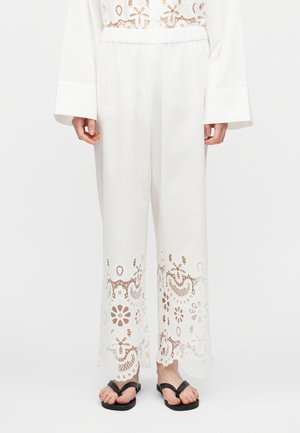 Fehér, bőszárú nadrág finom virágmintás kivágott csipkerészletekkel a szárvégeken, fekete papucsokkal viselve, egyszínű fehér háttér előtt.