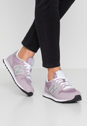 Osoba nosząca czarne spodnie i jasnofioletowe sneakersy New Balance 500 z białymi sznurówkami, stojąca na szarej powierzchni.