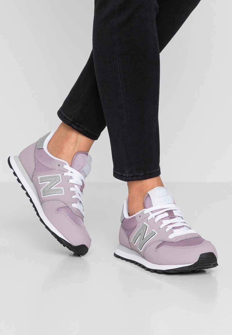 Osoba nosząca czarne spodnie i jasnofioletowe sneakersy New Balance 500 z białymi sznurówkami, stojąca na szarej powierzchni.