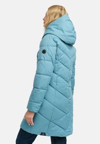 Turkusowa kurtka puchowa z dużym kapturem, wzorem rombów i czarną metką na rękawie. Ma prosty krój.