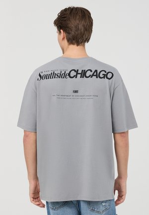 T-shirt in cotone grigio con una grande grafica nera sul retro, con testo e un piccolo elemento di design sottostante. Maniche corte e vestibilità rilassata.