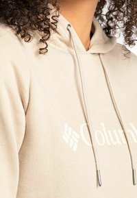 Sudadera beige con cordón, que presenta un logo blanco en el frente. Tejido suave y acabado texturizado con un corte holgado.