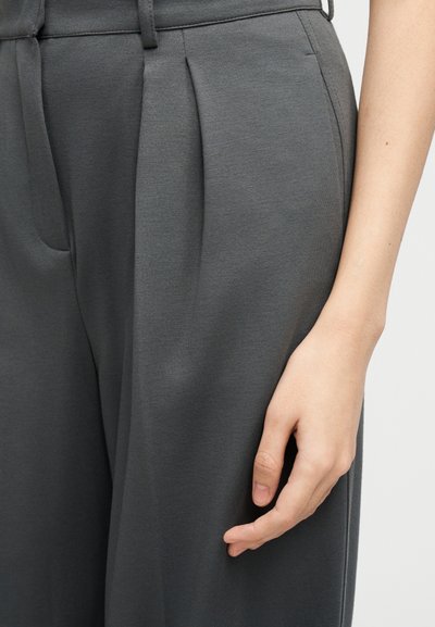 Κοντινή λήψη του χεριού ενός ατόμου που ακουμπάει δίπλα σε γκρι πτυχωτό παντελόνι με θηλιές για ζώνη και πλαϊνή τσέπη.