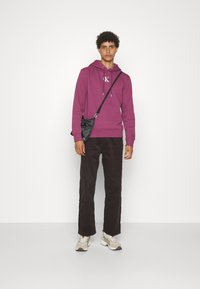 Felpa viola con logo, abbinata a pantaloni di velluto a coste scuri e sneakers chiare. Una borsa a tracolla nera aggiunge contrasto e funzionalità.