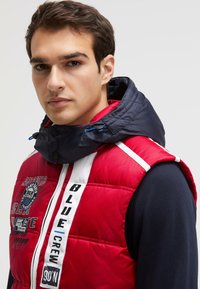 Red padded vest with a navy hood, featuring white stripes, logo details, and various text designs. Smooth texture with visible stitching.