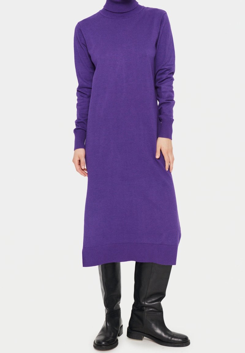 Femme portant une robe midi à col roulé à manches longues de couleur violette, associée à des bottes en cuir noires montantes jusqu'aux genoux, posant devant un fond blanc.