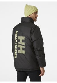 Černá péřová bunda s vysokým límcem, s velkým žluto-zeleným logem Helly Hansen na zádech. Nosená se světle zelenou pletenou čepicí.
