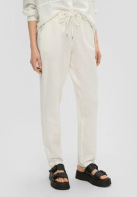 Białe lniane spodnie z elastycznym paskiem i bocznymi kieszeniami, w zestawieniu z czarnymi sandałami na platformie z podwójnymi klamrami.