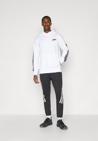 Sudadera blanca con capucha y logo, combinada con pantalones de chándal gris oscuro que tienen detalles laterales a rayas blancas. Zapatillas deportivas negras completan el conjunto.