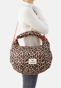 Person in einem weißen Oberteil, der eine große Schultertasche mit Leopardenmuster und gepolstertem Griff sowie einem Markenschild an der Seite trägt.