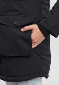 Veste noire isolante avec une finition texturée, dotée d'une poche à rabat fermée par un bouton noir, et d'un design de poignet ajusté.