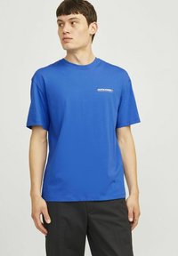 Mėlyna medvilninė marškinėliai trumpomis rankovėmis, apvaliu kaklu ir mažuoju baltu logotipu kairiajame krūtinės pusėje. Audinys atrodo lygus ir lengvas.