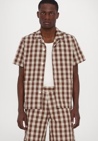 Brown and cream checkered short-sleeve shirt with a collar, buttons, and two front pockets, worn over a white tank top.