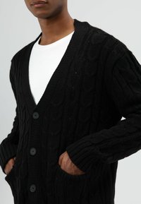 Zwarte kabelgebreide cardigan met een V-hals, geribbelde manchetten en zakken, voorzien van knoopsluitingen en een gestructureerd patroon.