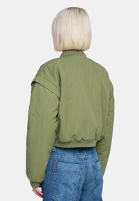 Olivgrön bomberjacka med ribbat krage och manschetter, som har en avslappnad passform och subtila axelsömmar. Bärs med blå denimjeans.