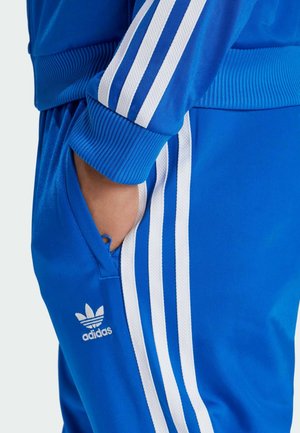 Mano en el bolsillo llevando un chándal azul de Adidas con tres rayas blancas en la manga y los pantalones, logo de Adidas en el bolsillo de los pantalones.
