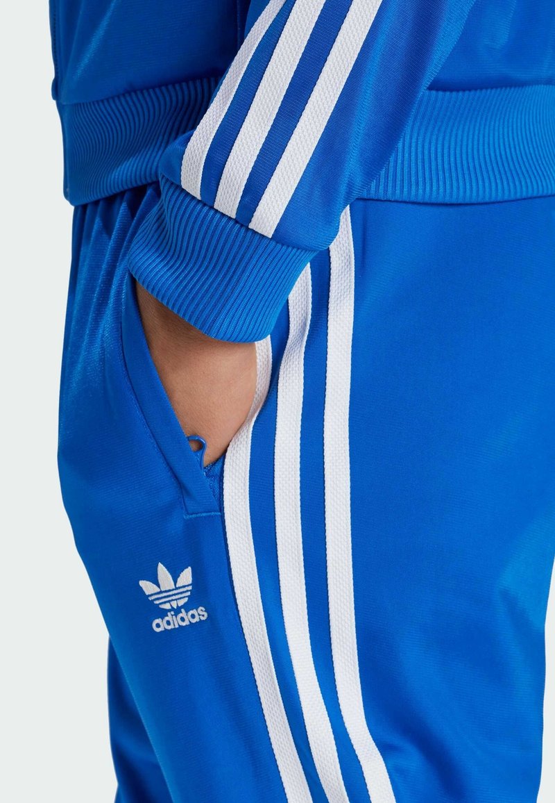 Ręka w kieszeni ubrana w niebieski dres Adidas z białymi trzema paskami na rękawie i spodniach, logo Adidas na kieszeni spodni.