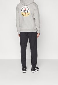 Harmaa huppari, jossa on valkoinen ja keltainen pyöreä Manchester Unitedin logo selässä, yhdistettynä mustiin verkkareihin ja mustiin urheilukenkiin.