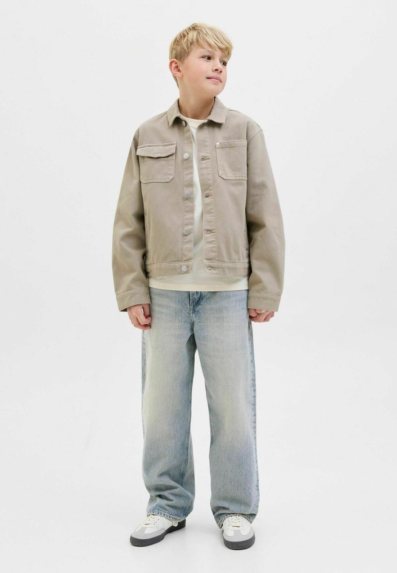 Veste beige clair avec deux poche poitrine, fermeture à boutons sur le devant et poignets côtelés, associée à un jean bleu clair à coupe ample et des baskets blanches.