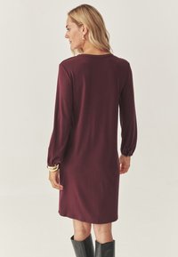 Femeie �îmbrăcată într-o rochie burgundă cu mânecă lungă și lungime până la genunchi, purtând un brățară aurie și cercei hoop, având pe picioare botine negre până la genunchi, întoarsă cu fața la noi.