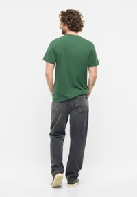 Dunkelgrünes Baumwoll-T-Shirt mit kurzen Ärmeln, kombiniert mit locker sitzenden, ausgewaschenen schwarzen Jeans und hellen Schuhen. Ansicht von hinten.