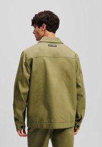 Olivengrøn jakke med krave, bagetiket og knapmanchetter. Fremstillet af et robust materiale med en glat tekstur og minimalistisk design.