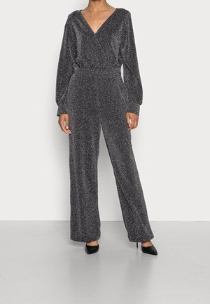 Combinaison noire avec un décolleté en V et des manches longues, présentant une texture scintillante, une taille cintrée et une silhouette à jambes larges. Associée à des talons noirs.