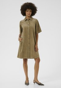 Femeie îmbrăcată cu o rochie din catifea bej, cu mâneci scurte și lungime până la genunchi, având un buton pe piept și purtând tocuri slingback cu imprimeu zebra, stând cu o mână în buzunar.