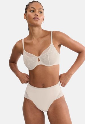 Femeie purtând un sutien bej din dantelă asortat cu chiloți cu talie înaltă, stând cu mâinile pe șolduri, pe un fundal simplu.
