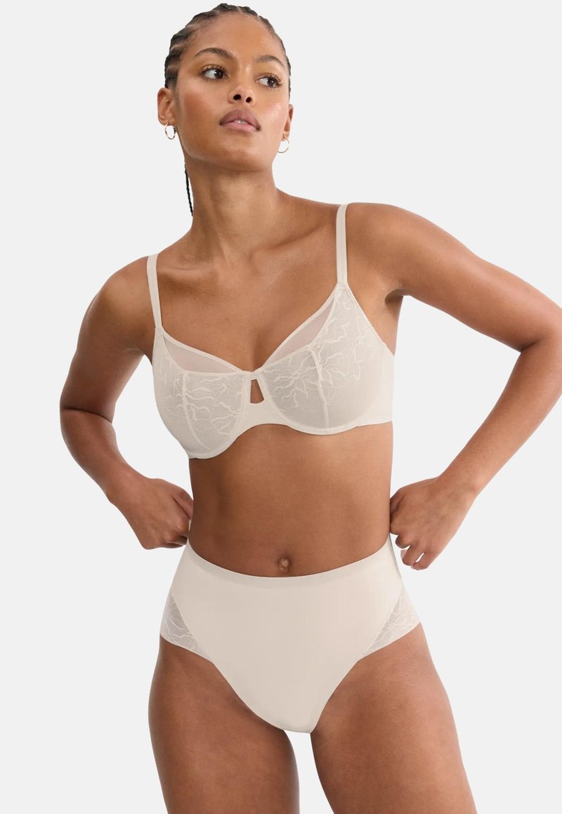Γυναίκα που φοράει σετ μπεζ δαντελένιο σουτιέν και εσώρουχο ψηλού μέσης, στέκεται με τα χέρια στη μέση μπροστά σε απλό φόντο.