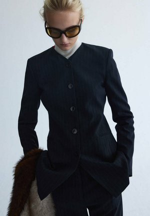 Person trägt einen dunklen Nadelstreifenanzug, weißen Rollkragenpullover und große braun getönte Sonnenbrille, hält ein braunes Pelzaccessoire.