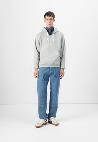 Sudadera gris con capucha, cordones blancos, pantalones de mezclilla azules con bolsillos y zapatillas blancas. Pañuelo atado alrededor del cuello.