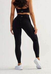 Leggings noirs avec une texture lisse, taille haute et logo en forme de cœur à l'arrière. Associés à un soutien-gorge de sport noir à bretelles et des baskets blanches.