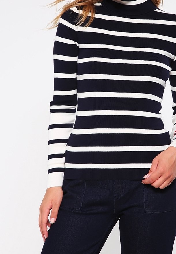 Femme portant un pull à col roulé rayé horizontalement noir et blanc à manches longues et un pantalon foncé, debout avec une main près de sa hanche.