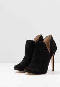 Musta mokkanahka avonaisella varpaalla varustetut nilkkurit, joissa on korkea stiletto-korko ja leikattu muotoilu. Sileä pinta ja minimalistinen siluetti.
