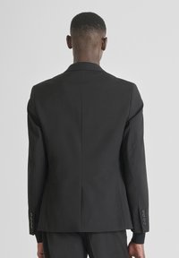 Muškarac u uskom crnom odijelu i hlačama, prikazan s leđa na jednostavnoj svijetloj pozadini.