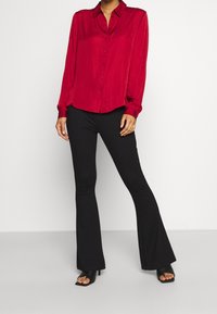 Červená košile na knoflíky s dlouhými rukávy a límečkem, doplněná černými rozšířenými kalhotami a černými sandály na otevřenou špičku. Hladká, lehká látka.