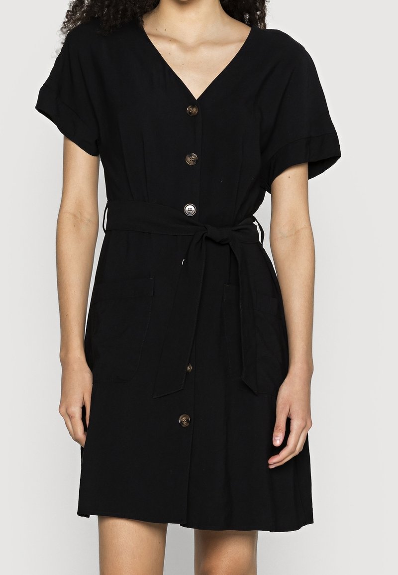 Femme portant une robe noire boutonnée jusqu'aux genoux, manches courtes et ceinture à nouer à la taille.