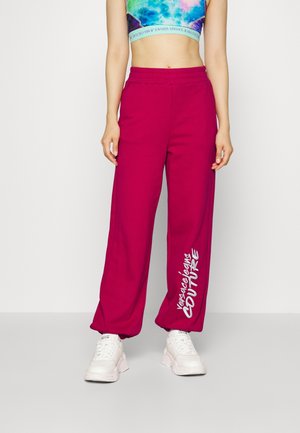 Personne portant un pantalon large magenta avec le texte blanc "Versace Jeans Couture" et des baskets blanches épaisses, visible du milieu du torse jusqu'aux pieds.