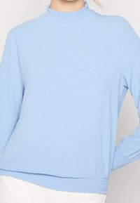 Maglietta a maniche lunghe blu chiaro con un collo alto arricciato. Tessuto testurizzato con una vestibilità ampia e dettagli arricciati nei polsini e nell'orlo.