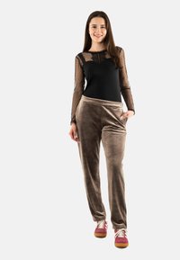 Body negro con mangas transparentes con puntos, combinado con pantalones de terciopelo color beige y zapatillas multicolores. La modelo se encuentra de pie en el centro, con las manos en las caderas.