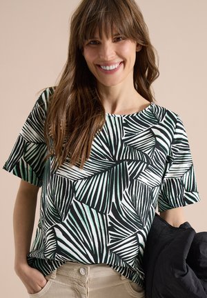 Smilende kvinde med langt brunt hår iført en skjorte med sort, hvidt og grønt bladmønster og holder en sort jakke i den ene hånd.