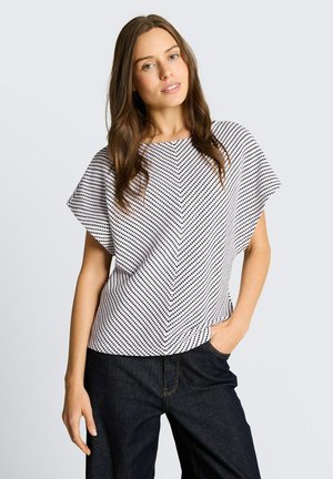 Młoda kobieta o długich brązowych włosach, ubrana w biało-czarny wzorzysty top z krótkim rękawem i ciemne dżinsy, stoi z jedną ręką w kieszeni.