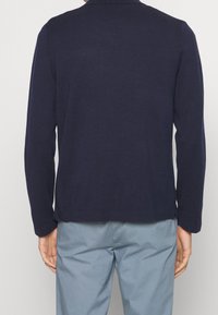 Man som bär en marinblå långärmad skjorta och ljusblå byxor, sedd bakifrån mot en enkel bakgrund.