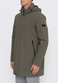 Giacca impermeabile verde oliva con collo alto e cappuccio regolabile, con cerniera frontale e tasche laterali con zip. Logo nero sulla manica.