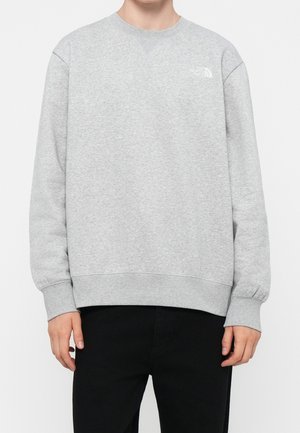 Person iført lysegrå crewneck-sweatshirt med lille hvidt The North Face-logo på venstre bryst og sorte bukser.