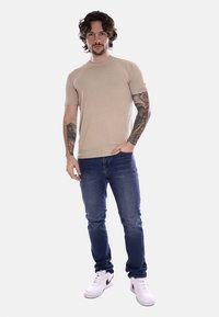 T-shirt beige a maniche corte, design aderente, indossato con jeans blu scuro. Sneakers bianche con accenti neri. Modello in piedi contro uno sfondo neutro.