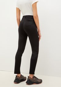 Žena oblečená v lesklých černých slim-fit kalhotách, bílém tričku s krátkým rukávem a černých sportovních botách s leopardím potiskem stojí proti bílé zdi.