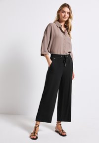 Collared, light brown shirt with rolled sleeves, paired with black wide-leg trousers featuring an elastic waist and side pockets. Black sandals.