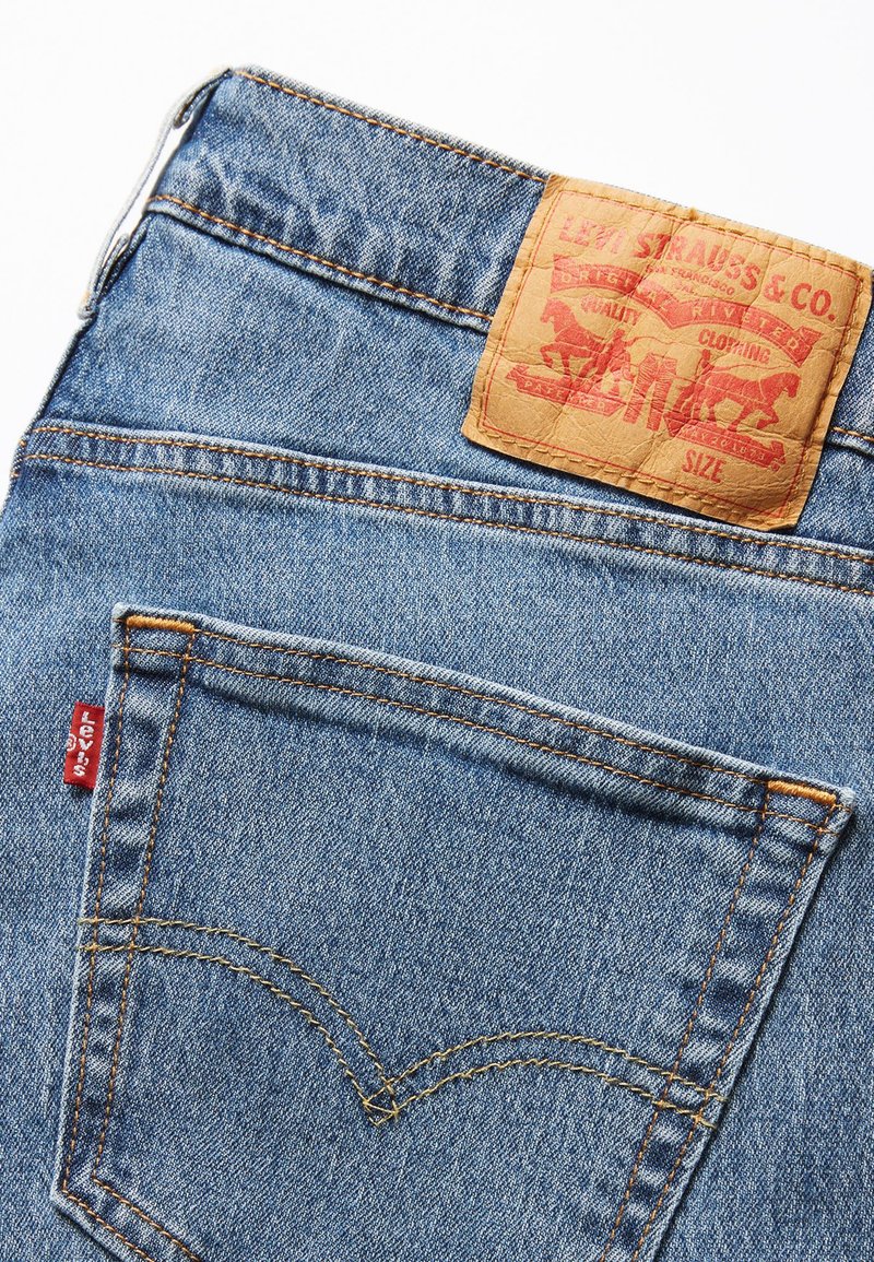 Jeans de mezclilla azul con un parche de cuero marrón etiquetado "Levi Strauss & Co." en la cintura, con costuras naranjas y una pequeña etiqueta roja.